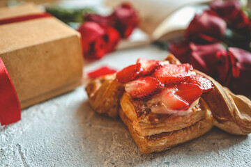Delicious puff pastry topped with fresh strawberries placed beside a kraft gift box tied with a red ribbon, surrounded by vibrant red roses and petals on a textured light surface