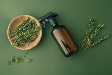 Flat lay of fresh rosemary sprigs in a wooden bowl with an amber spray bottle on a green textured...