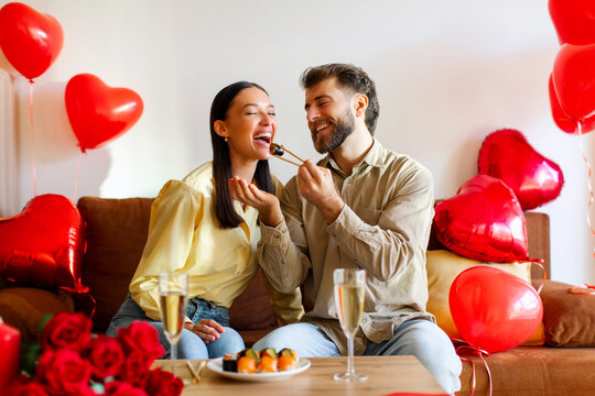 Take it. Couple celebrating Valentine's day at home, man feeding wife with sushi rolls, sitting in decorated room among heart shaped balloons - Powered by Adobe