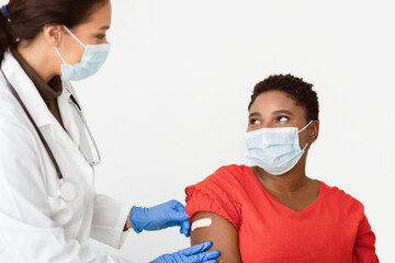 Covid-19 Vaccination. Black Woman Getting Vaccinated Against Coronavirus, Doctor Sticking Adhesive Plaster Strip After Corona Virus Vaccine Injection Shot On White Studio Background, Wearing Face Mask