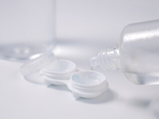 Transparent liquid dripping into an open contact lens case from a plastic solution bottle on a white background. Concept of lens care, hygiene, and eye health