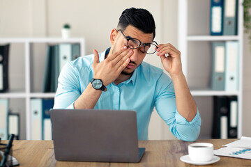 Young Arab businessman having tired irritated eyes, sitting at desk in front of laptop, exhausted...