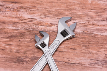Two adjustable wrenches lie crossed in an X shape on a wooden surface. Silver metal and black handles contrast sharply with the natural wood, highlighting their functionality