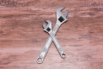 Two adjustable wrenches lie crossed in an X shape on a wooden surface. Silver metal and black handles contrast sharply with the natural wood, highlighting their functionality