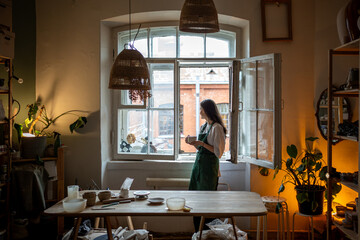 Thoughtful woman potter drinks tea and looks out window in workshop. Female taking coffee break on work, beginning of working day, artisan routine, creative occupation, slow living of woman ceramist