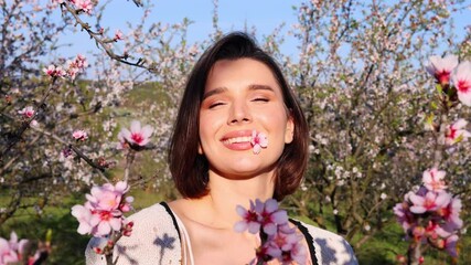 beautiful woman in almond trees orchard. girl with flowers in mouth or blowing petals from palm.female walking on grass, close up of hand touching pink flowers.smiling woman kissed by sun rays.