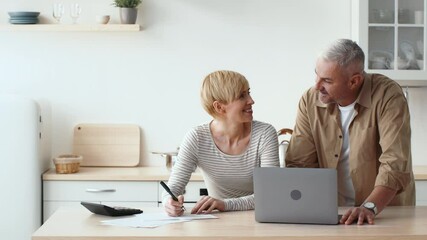 Two individuals engage in collaborative work at a light-filled kitchen table, with one person writing on paper while the other observes from a laptop. The atmosphere is casual and productive. - Powered by Adobe