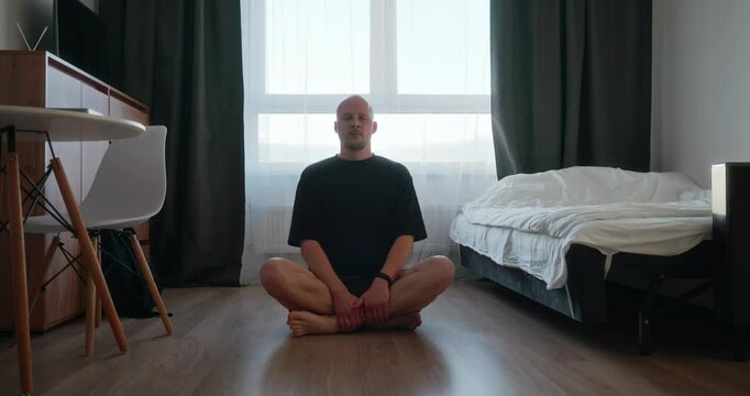 A single bald millennial man in a black T-shirt is sitting on the floor of a bedroom in an empty apartment and meditating, doing breathing exercises. Morning of a single man in a minimalist apartment