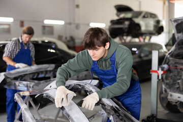 Young professional auto body technician in blue overalls and casual green hoodie, engaged in...