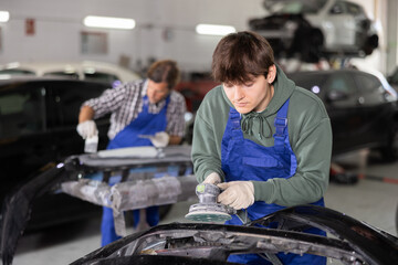 Young guy mechanic in uniform polishes part of car with machine in car service station