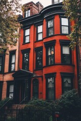 Fototapeta premium A charming bright red brick townhouse stands out beautifully among the lush, leafy green trees, set against the backdrop of an overcast urban neighborhood