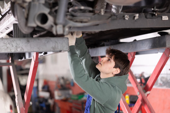Skilled focused young mechanic in warm olive hoodie and blue overalls engaged in repair of undercarriage of vehicle raised on lift in auto repair workshop - Powered by Adobe