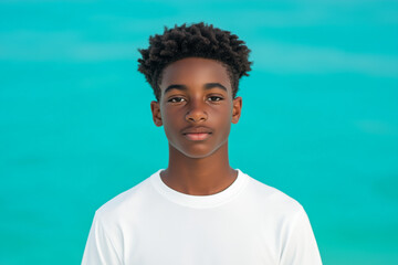 Calm caribbean teenage boy in his early teens wearing a white shirt and standing against a bright turquoise background