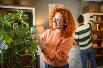young adult smiling woman take care and spray with water home plant