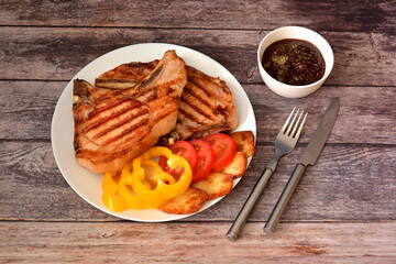 Grilled pork entrecote with tomatoes, sweet peppers and potatoes on a wooden table, next to a cup with sauce and cutlery.