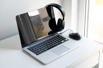 Open silver laptop, black wireless  headphones and black computer mouse on a white desk