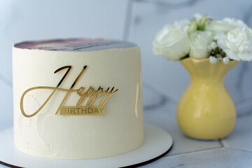 Elegant minimalist white birthday cake with golden Happy Birthday topper on cake board, placed next to a yellow vase