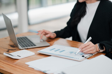 Professional woman reviewing charts and graphs on laptop and documents at a wooden desk, focusing on financial analysis.
