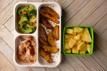 bento set lunch box. potato wedges, chicken steak, and stir-fried broccoli carrots. healthy low-fat food. sliced pineapples. 