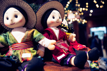 Two traditional Ecuadorian dolls wearing colorful clothing and sombreros, sitting on a shelf against a blurred background of lights.