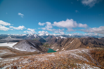 Sunny top view to most beautiful alpine lake between stony hill in freshly fallen snow and sharp rocky ridge in sunlight. Large colorful mountain range with snow-white peaks in far away in low clouds.
