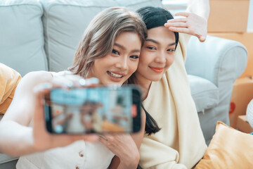 Fototapeta premium Two young Asian women smiling and happy while taking a selfie in their living room at home, looking at the camera
