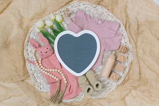 A delicate and feminine pregnancy announcement setup featuring soft textures, pastel pink tones, and spring elements. Includes a baby girl onesie, wooden toys, knit booties, and tulips arranged on a c