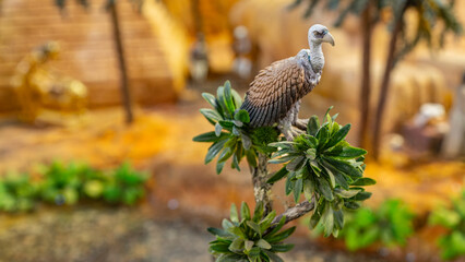 Close-up of a realistic vulture figurine perched on a branch, with a blurred natural background resembling a desert scene.