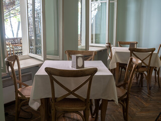 Empty vintage wood dining table set with white tablecloth and wooden chair seats on rubber tiles floor near windows, relaxation bright dining room background with nobody.