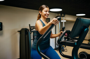 Smiling Woman Exercising on Elliptical Machine in Modern Gym Setting. She is wearing blue workout clothes.