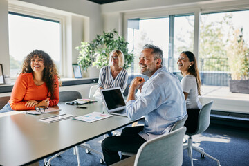 Smiling Team Enjoying a Productive Meeting in a Bright Office Space. They all look smiling at a colleague standing at the head of the table presenting a project.