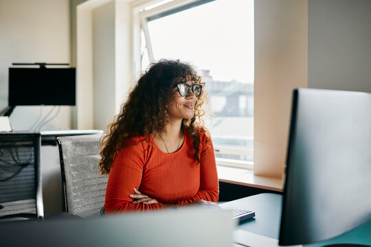 Confident Woman Working at Her Desk in a Modern Office Setting. She is smiling while wearing an orange sweater and glasses. Her hair is long and curly.