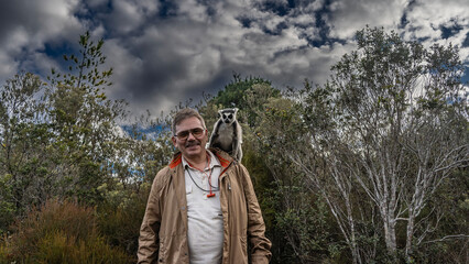 Ring-tailed lemur catta sits on a man's shoulder, looking at the camera. The man smiles happily. Behind them, the trees are against the blue sky and clouds. Madagascar. Lemur Island. Nosy Soa Park © Вера 