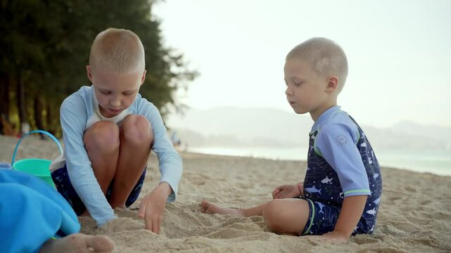 Two young brothers sitting close together, building sandcastle and playing on golden sandy beach, sharing joyful moment during sunny summer vacation