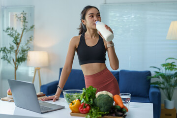 Young asian sportswoman drinking milk after training at home with fresh vegetables and laptop on the kitchen table, promoting a healthy lifestyle combining fitness and balanced nutrition