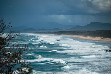 A dramatic coastline with waves crashing onto the sandy shore under a moody sky. The ocean's vibrant blues contrast with the dark clouds, creating a powerful and dynamic atmosphere. In the distance, h