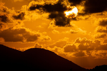 A mesmerizing sunset paints the sky in vivid hues of orange and gold, with dramatic clouds casting intricate shadows across the scene. The silhouette of distant hills adds depth, while the reflective 