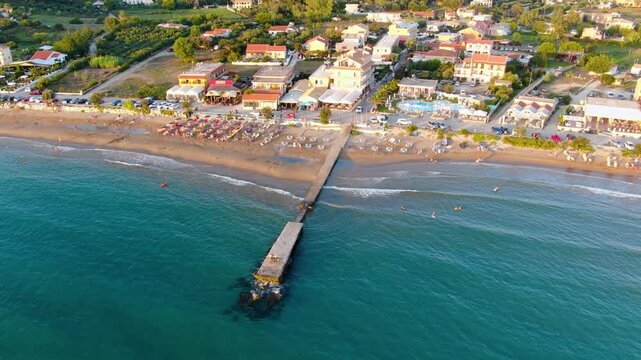 a pier extending into turquoise water, meeting a sandy beach with beach umbrellas.Arillas beach in Corfu island, Greece