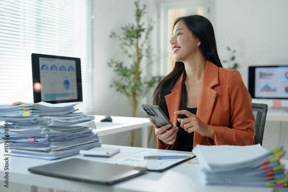 Poster smiling asian businesswoman using her smartphone while working with documents at a modern office des