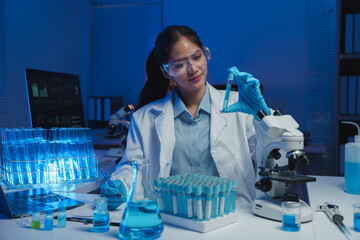 Female scientist wearing lab coat and gloves analyzing test tube with blue liquid, taking notes and...