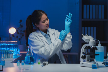 Female scientist carefully putting on blue nitrile gloves, preparing for experiment in modern laboratory with test tubes, flasks, and microscope under blue light