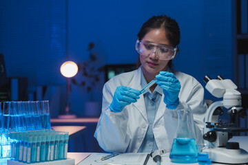 Female scientist wearing lab coat and protective glasses holding test tube with blue liquid while...