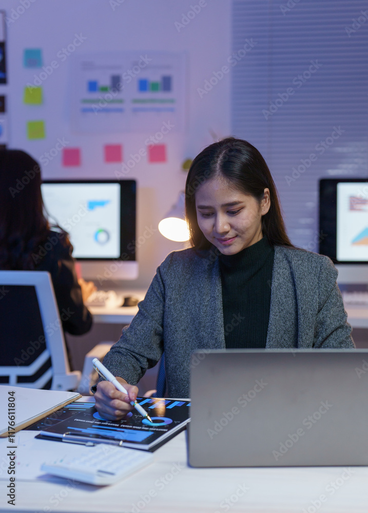 Poster young professional woman using laptop and reviewing marketing data working late in modern office wit