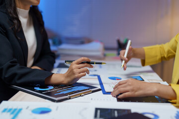 Two businesswomen are analyzing financial data, using pens to point at charts and graphs while discussing project strategy and investment opportunities
