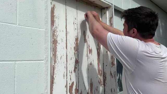 Caucasian man in a white t-shirt scraping old flaking paint off a wooden door set into a concrete block structure with a metal scraper tool. 