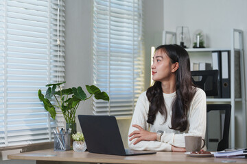 Young asian businesswoman sitting at her desk, working on a laptop and gazing thoughtfully out the window during a peaceful break in a modern office environment