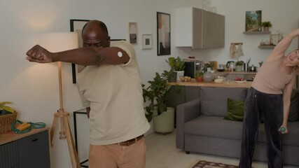 Medium shot of mid-aged African American man with CGM on his arm and Caucasian woman with light weight dumbbells training together in cozy living room