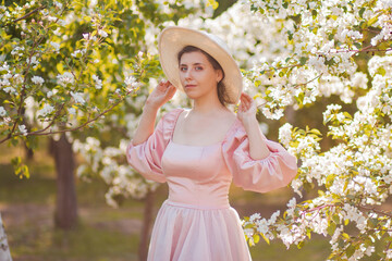 Portrait of a young woman wearing a pink dress and hat posing against the backdrop of a spring park and white blooming apple trees.