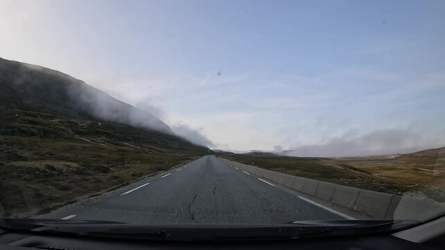 Driving through the tundra in northern Norway on E69 road. Grasslands and a rocky hill surround the area. Low clouds create a serene and atmospheric view of the Arctic landscape.