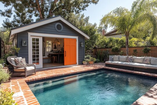 Small barn-style gray and white building with orange door next to in-ground pool on red brick patio, California backyard, daytime.